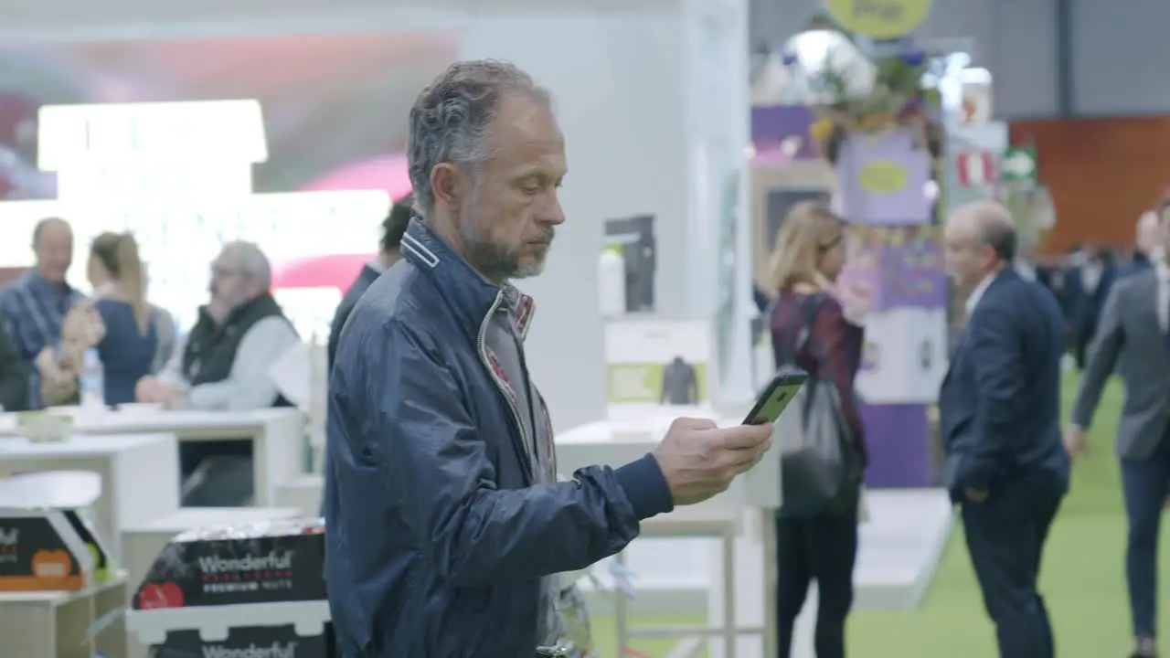 Man standing around looking down at phone busy conference people in background