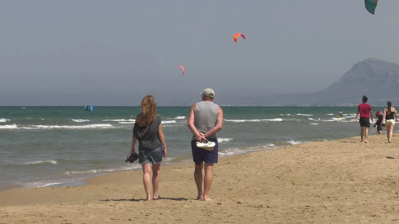 Middle aged couple walking on sandy beach with kitesurfers in background wide shot