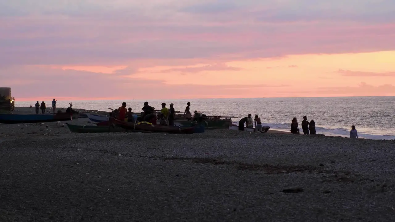 People relaxing and socialising near fishing boats on Beto Tasi beach during pink sunset in capital Dili Timor-Leste Southeast Asia