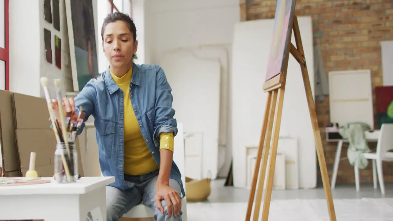 Biracial woman selects paintbrushes in a bright studio