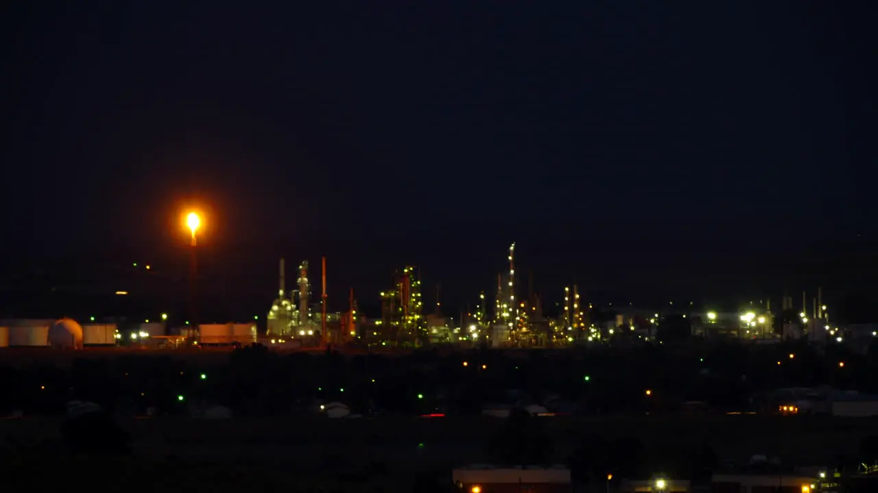 Twilight falls on an oil refinery with flaming tower and lights