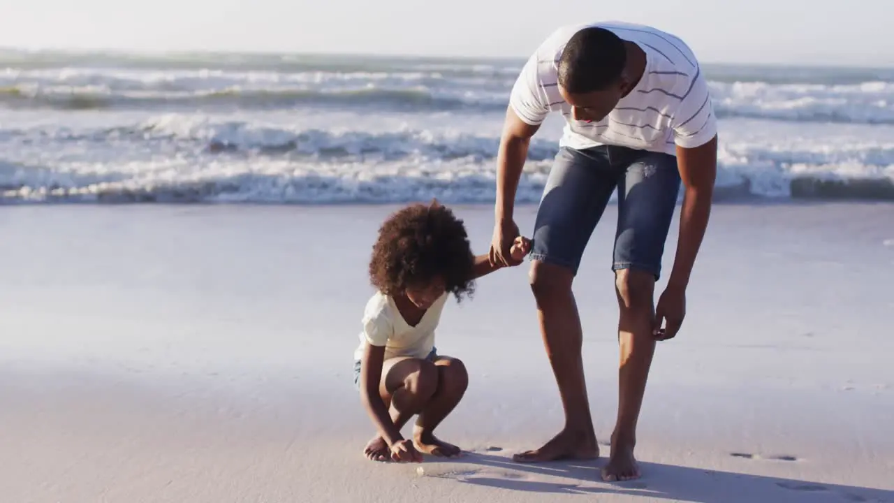 African american father and daughter picking up shells from the sand together at the beach