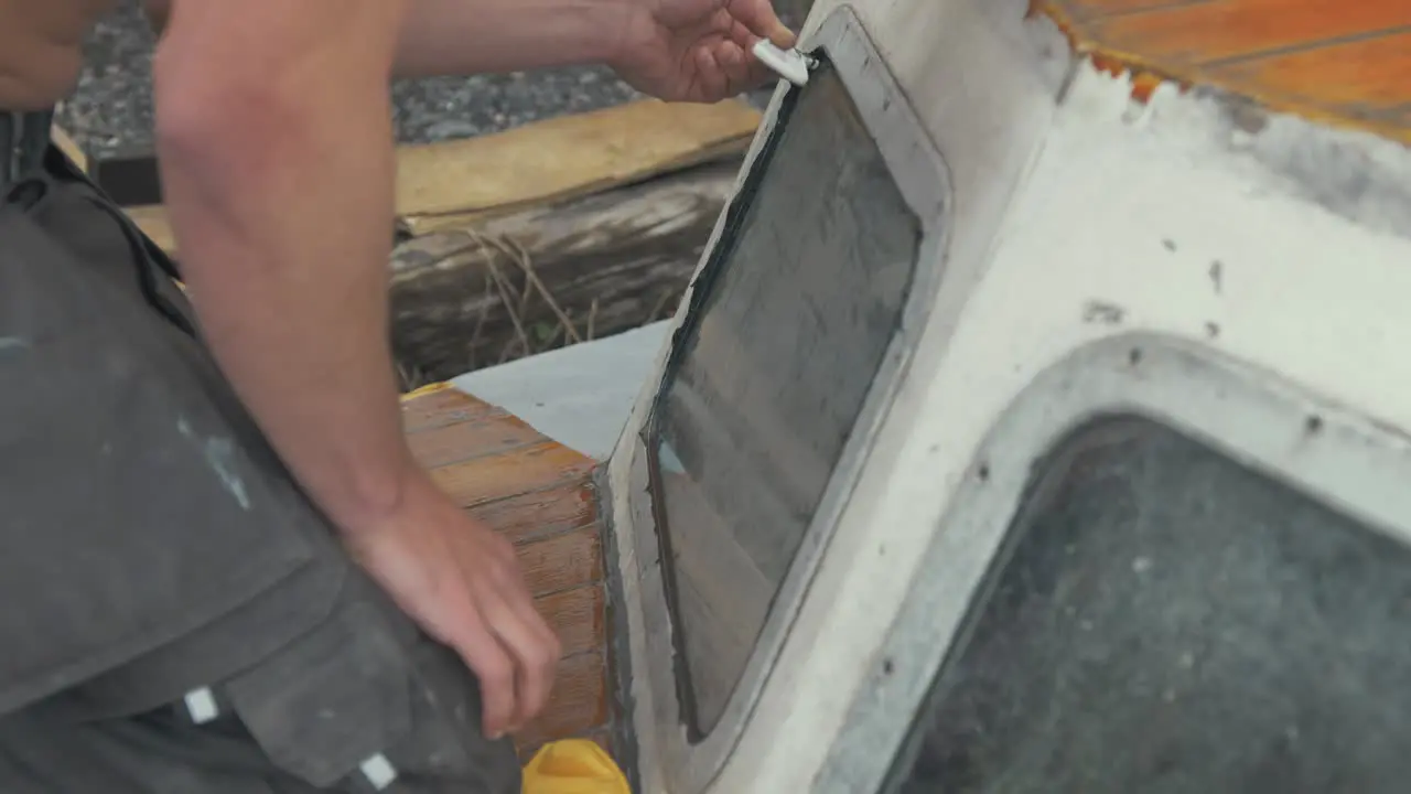 Young carpenter using scraper to flatten sika flex mastic sealant on windows on bow of wooden boat