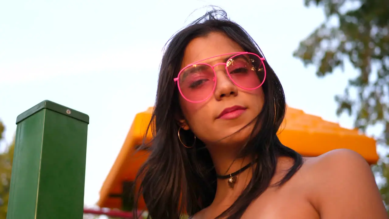 A cute hispanic woman hipster wearing retro pink sunglasses in a park playground SLOW MOTION