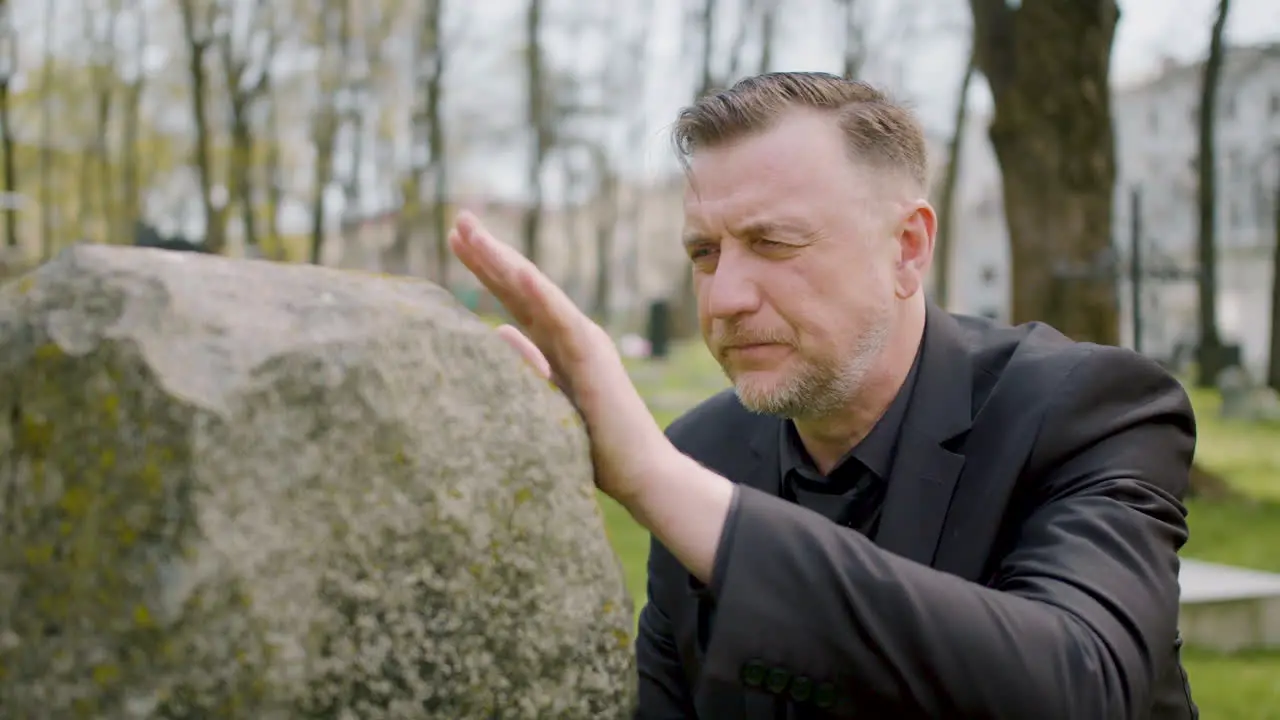 Close Up View Of Man In Black Suit Kneeling In Front Of A Tombstone And Touching It