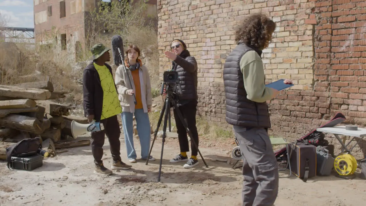 Three Production Coworkers Talking About A Recording Project And Laughing In Front Of A Camera On A Tripod In The Street While Another Worker Taking Notes 1