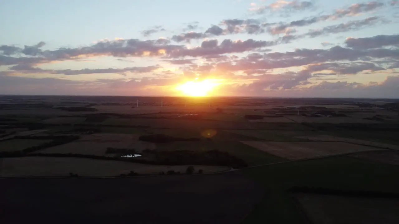 Drone flies away from sunset with lush green farm landscape on the ground in denmark