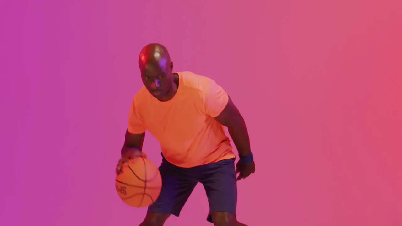 Video of african american male basketball player bouncing ball on pink to orange background