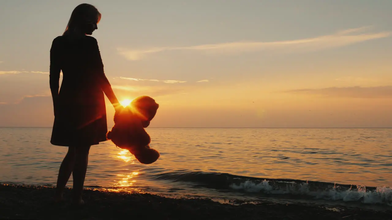 A Woman With A Teddy Bear In Hand Is Standing On The Seashore When The Sun Sets Childhood Dreams