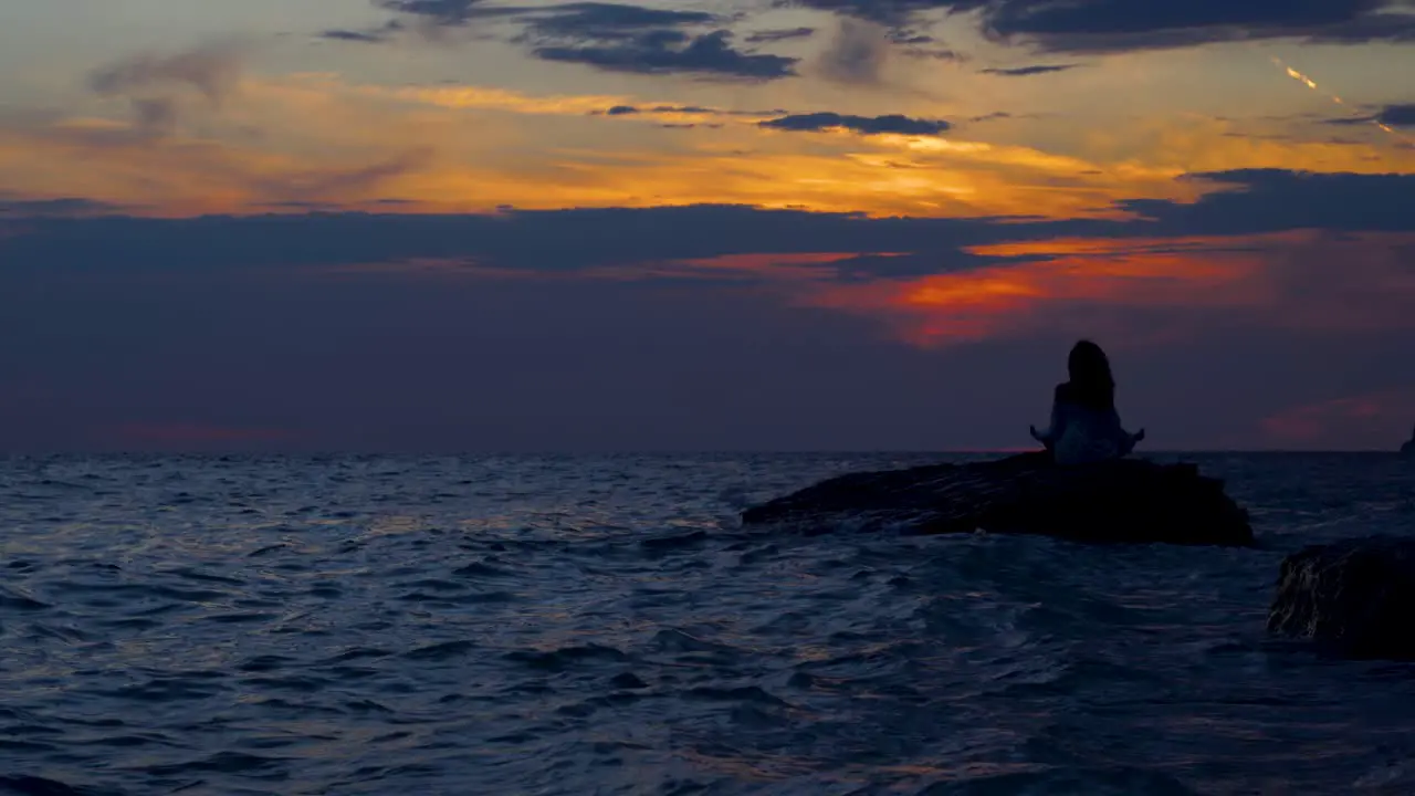 Woman sitting on rock surrounded by dark sea water at beautiful sunset doing Yoga exercises