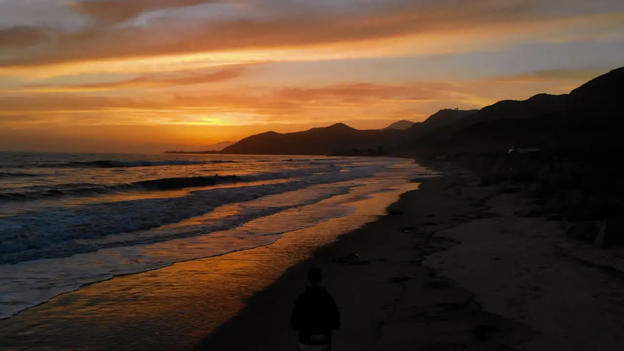 AERIAL drone shot off of the California coast at sunset with bright orange sky and person silhouette on the beach