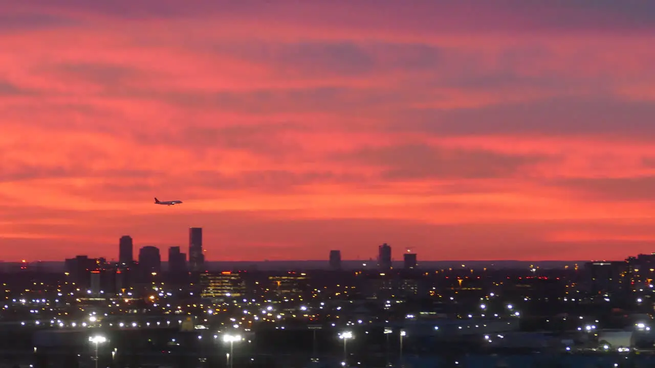 Silhouette of plane landing above dusk cityscape with glowing lights and red sunset sky