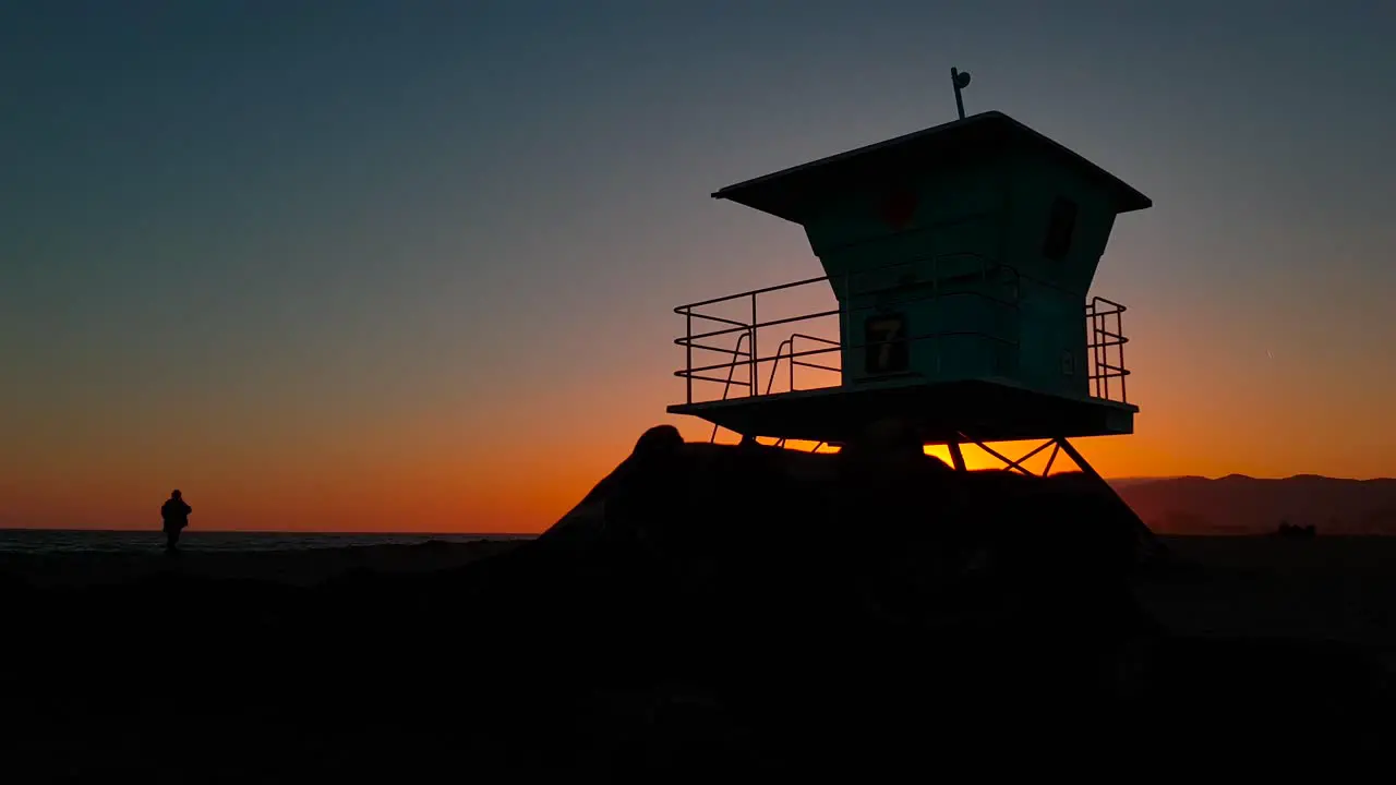 Slow and low sideways sunset shot of Lifeguard house  tower with silhouettes of people by the shore at sunset at San Buenaventura State Beach in Ventura California United States