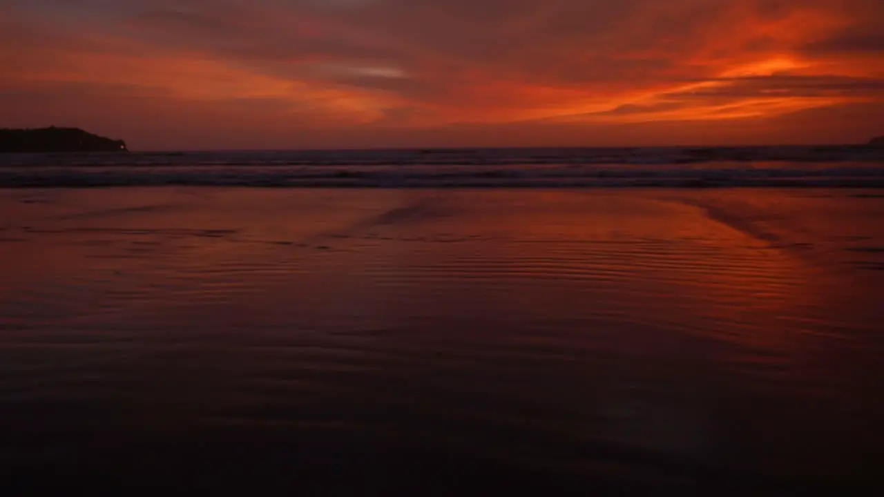 Panoramic seascape view of ocean waves crashing on the sandy coastline during a bright red and orange sunset on a cloudy evening