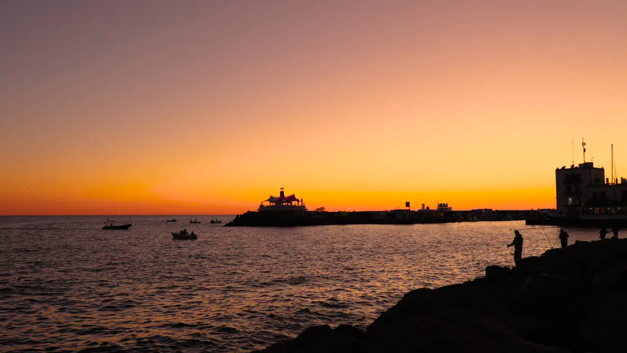 Fisherman silhouette on stony coast during orange sunset casting fishing line on open sea in background is city and harbor of sunset Grand Canary island valley 4k slow motion capture at 60fps