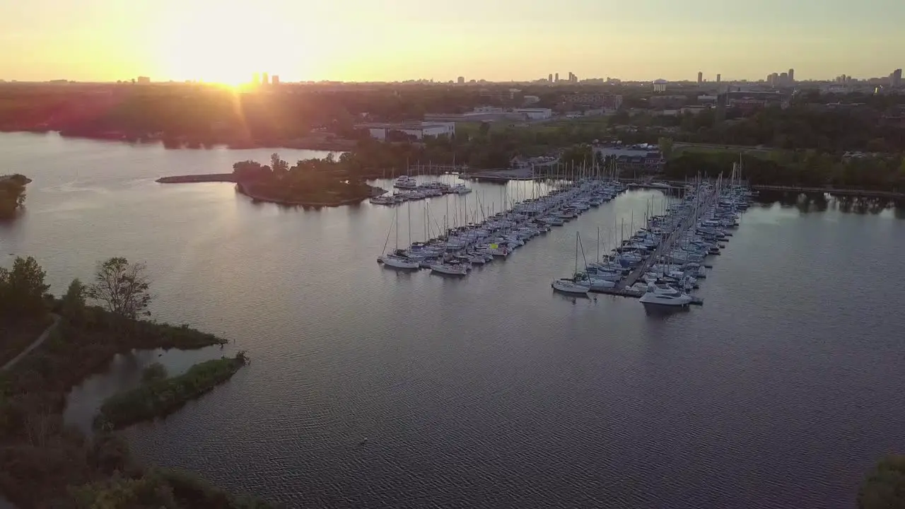 Aerial Sunset Wide Shot Flying Toward Sailboat Marina Yacht Club Dock In Lake Bay Among Green Trees And Close Birds With City Buildings Skyline In Background In Toronto Ontario Canada