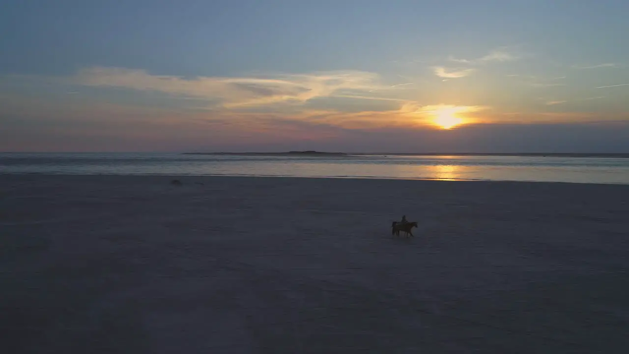 Aerial orbit drone shot around a man and a woman riding horses on a beach while waves hit the coast on a warm and sunny evening