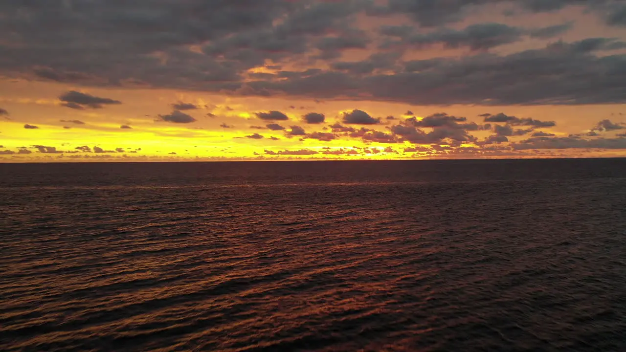 Aerial flight backwards over calm ocean with a dramatic bright yellow orange and purple sunset