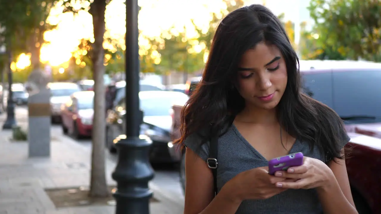 A young woman walking on an urban city street sidewalk texting or using social media on her smartphone at sunset SLOW MOTION