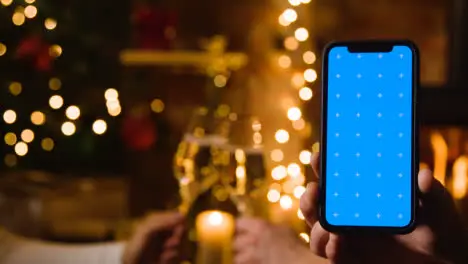 Couple Making Christmas Toast With Champagne In Front Of Fire At Home With Blue Screen Mobile Phone