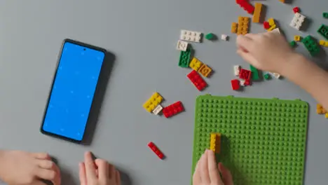 Overhead Shot Of Two Children Playing With Plastic Construction Bricks Next To Blue Screen Mobile Phone