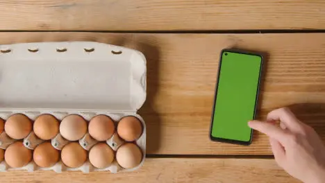 Overhead Shot Of Person Following Recipe On Green Screen Mobile Phone With Box Of Eggs On Wooden Table