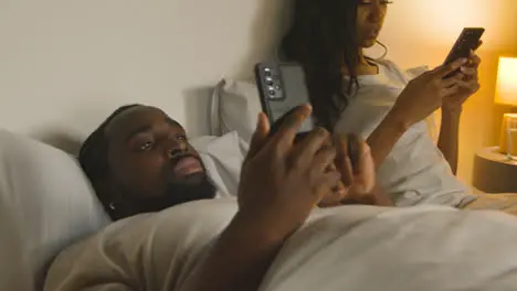 Couple With Relationship Problems At Home At Night Lying In Bed Both Looking At Mobile Phones 1