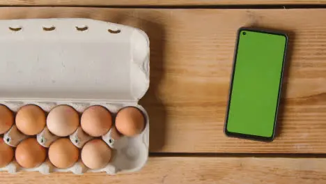 Overhead Shot Of Person Choosing Egg From Cardboard Box On Wooden Table With Mobile Phone