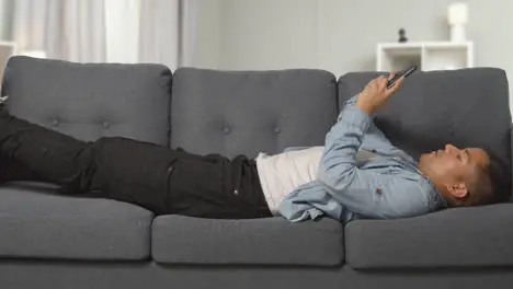 Young Man Lying On Sofa At Home Scrolling Through Mobile Phone