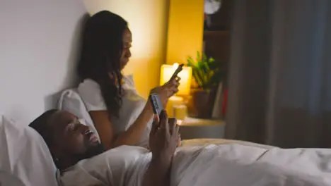 Couple With Relationship Problems At Home At Night Both Looking At Mobile Phones In Bedroom 11