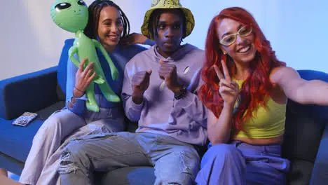 Studio Shot Of Group Of Gen Z Friends Sitting On Sofa Posing For Selfie With Toy Alien On Mobile Phone