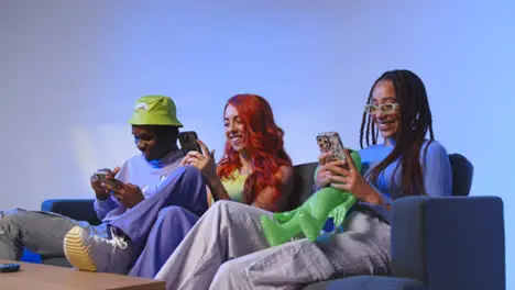 Studio Shot Of Group Of Young Gen Z Friends Sitting On Sofa Gaming And Using Social Media On Mobile Phones