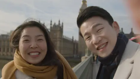 Young Asian Couple On Holiday Making Video Call Home On Phone At Houses Of Parliament In London UK