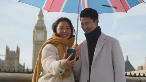 Young Asian Couple On Holiday Posing For Selfie In Front Of Houses Of Parliament In London UK With Umbrella 1