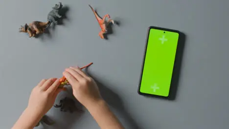 Overhead Shot Of Boy Playing With Toy Dinosaurs Next To Green Screen Mobile Phone