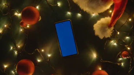 Overhead Shot Of Revolving Blue Screen Mobile Phone With Christmas Decorations Lights And Santa Hat