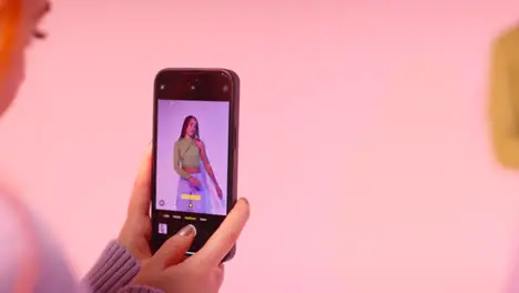 Studio Shot Of Woman Taking Photo Of Friend Dancing On Mobile Phone Against Pink Background