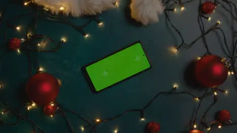 Overhead Shot Of Revolving Green Screen Mobile Phone With Christmas Decorations Lights And Santa Hat 