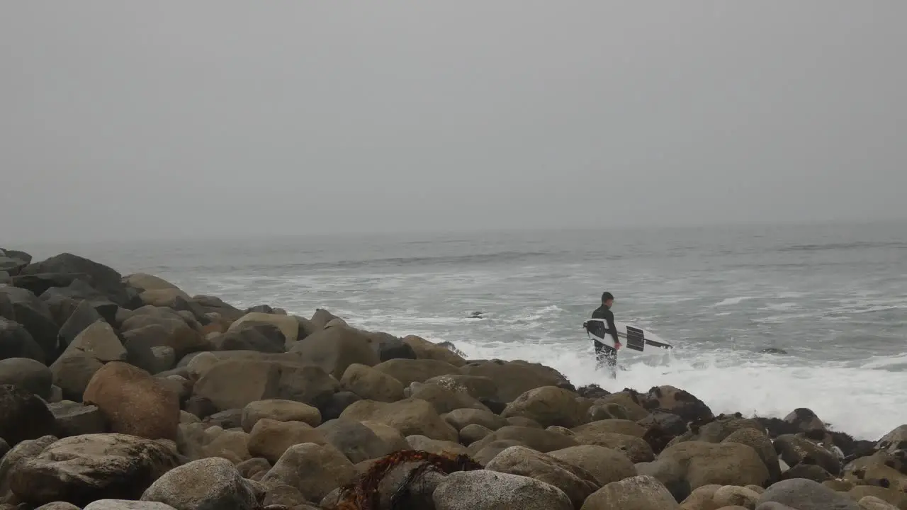 Surfer standing on rocky ocean shore as he gets ready to paddle out to sea