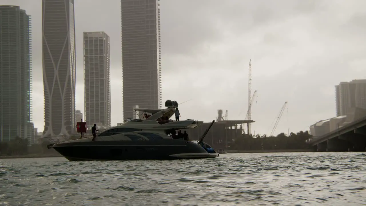 Yacht in a harbor with a man walking along the edge of it with a city filled with skyscrapers under a cloudy grey sky in the background