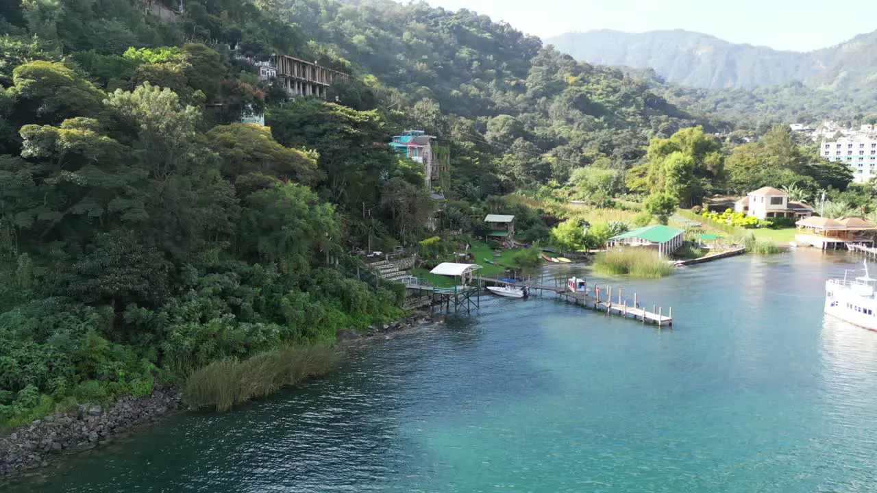 Drone view in Guatemala flying over a blue lake next to a green mountain with a wooden walkway and a hotel on a hill on a sunny day in Atitlan