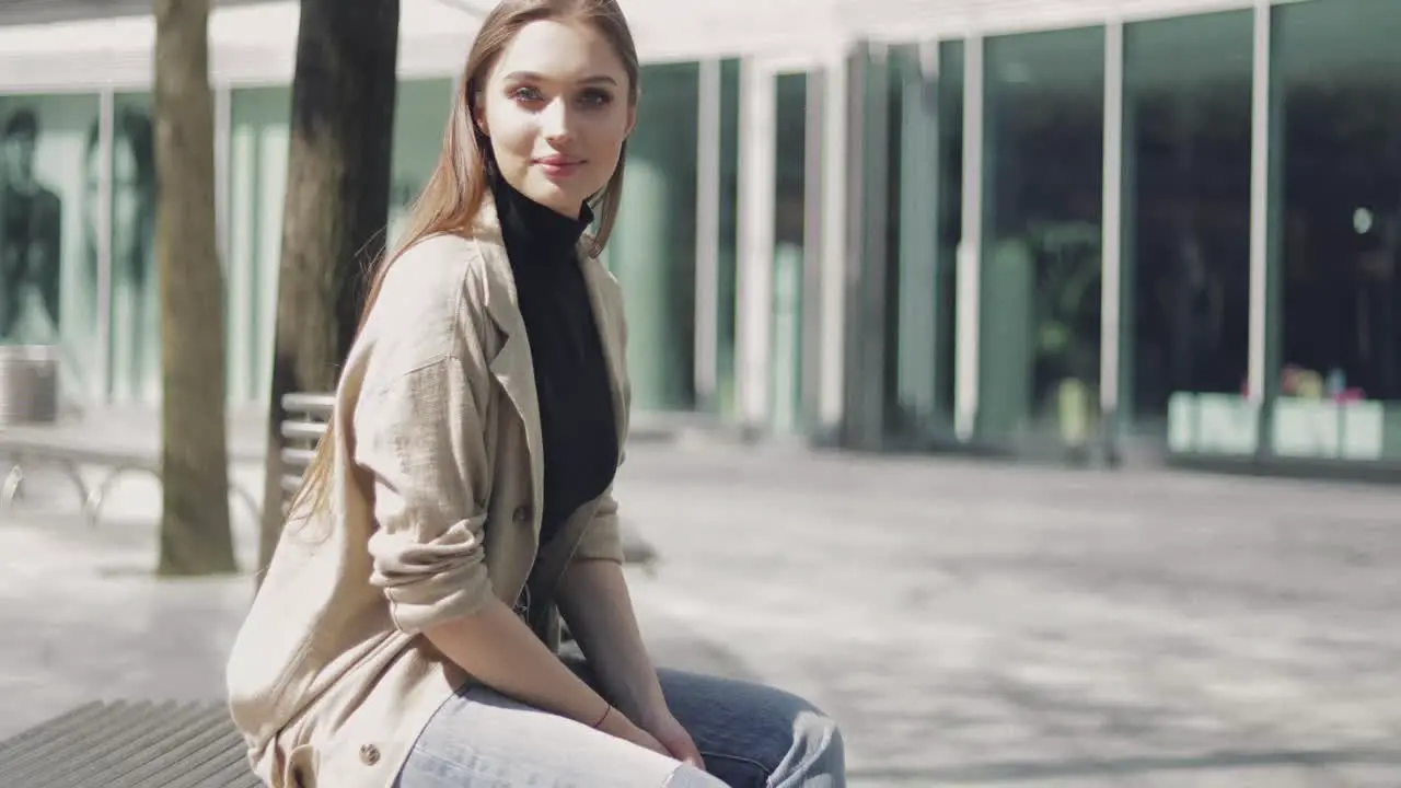 Smiling beautiful female in coat sitting on bench and looking at camera