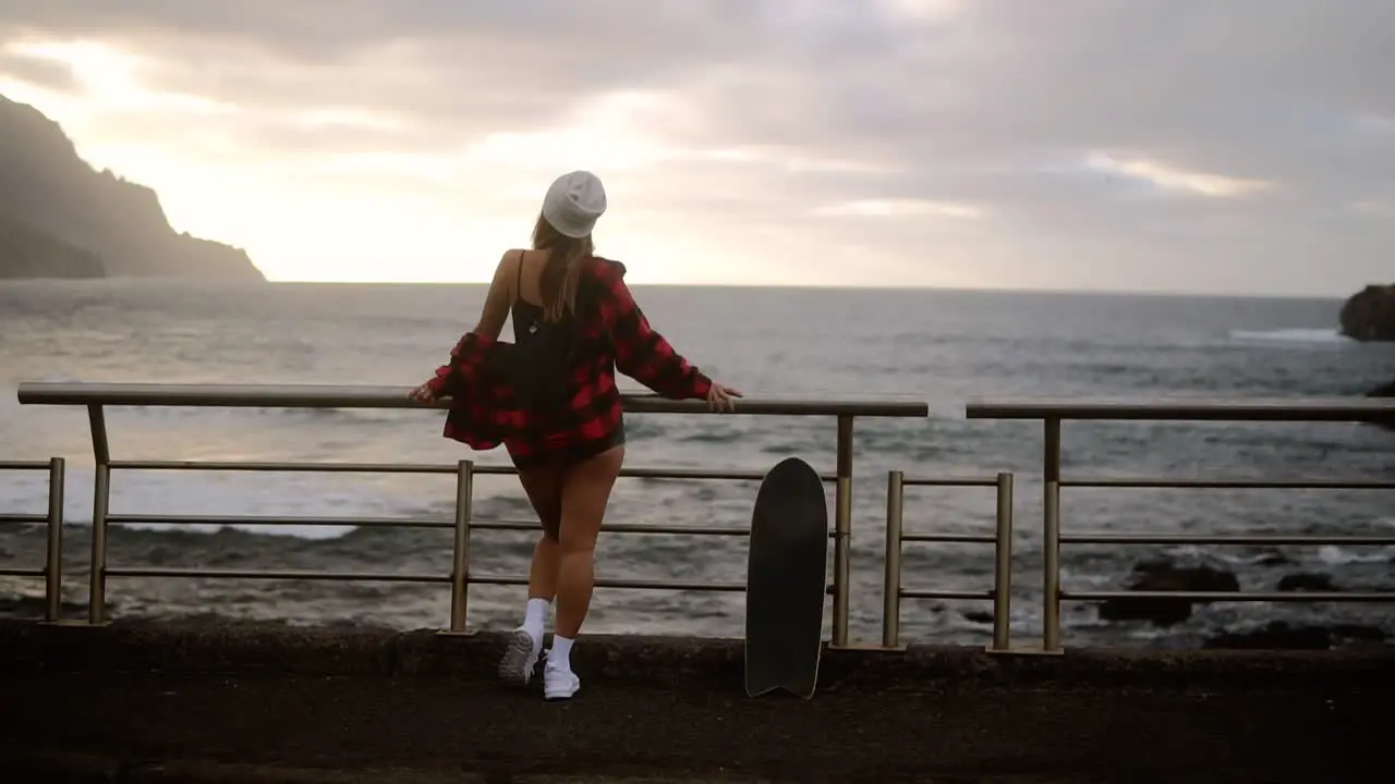 Full Length Sporty Builded Woman In Hat Plaid Coat And White Sneakers Enjoying Time By The Seaside On A Cloudy Day At Sunset Standing Leaning On Railings With Skateboard Watching Stormy Ocean From Promenade