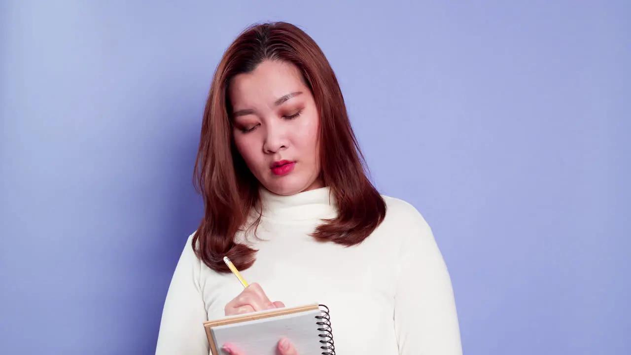 Happy beautiful Asian girl making notes in a paper notebook writing creative ideas and to-do list indoor studio shot isolated on purple background