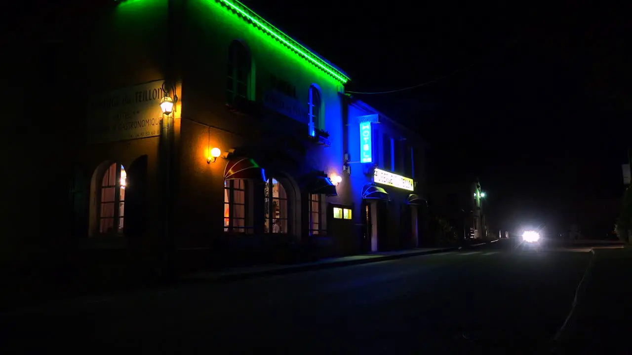 A car passes a small French hotel at night with neon sign flashing