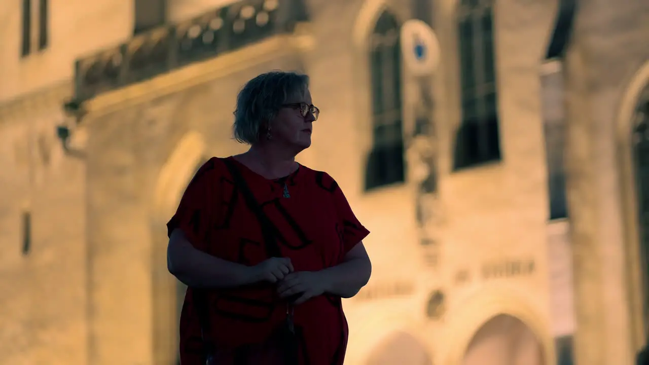 Older white Woman with red T-shirt and glasses standing and looking around in front of illuminated historical European church at night in slow motion