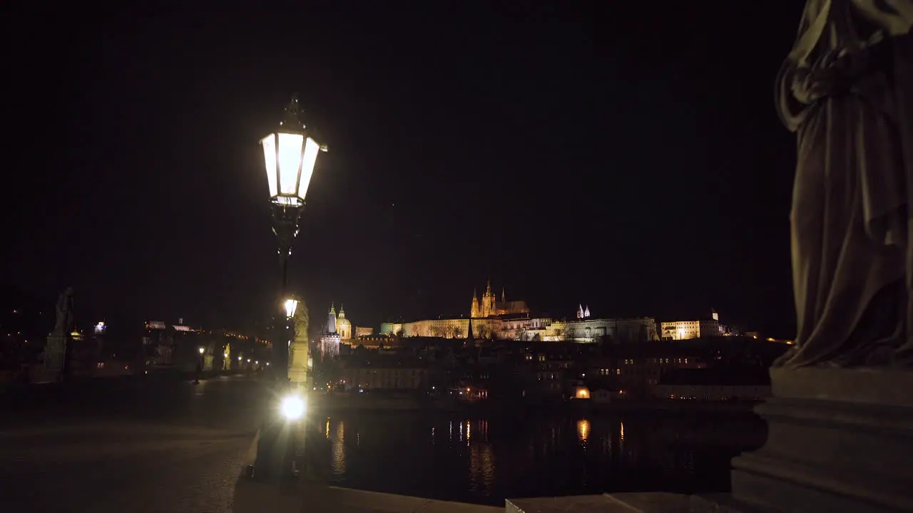 An ornate stone cross with crucified Jesus on Charles bridge in Prague at night during a Covid-19 lockdown statues and lanterns on both sides of it lit Prague castle in the background 4k shot