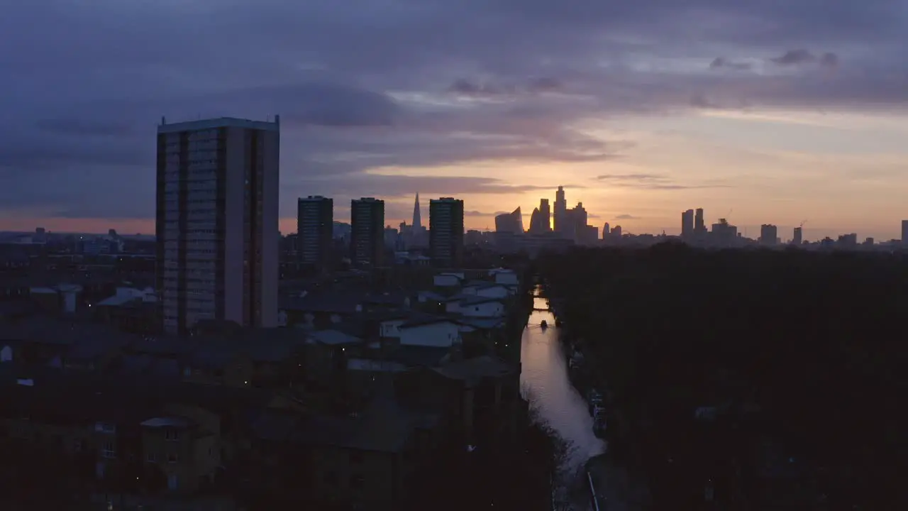 Low dolly back Aerial drone shot of house boat on London Canal Victoria park towards city skyline to highway at sunset