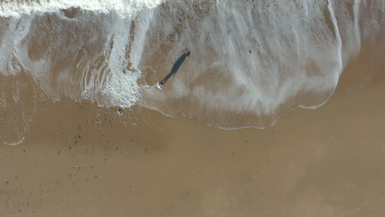 Aerial shot following a person walking on an empty beach with white foam waves crashing at he's feet