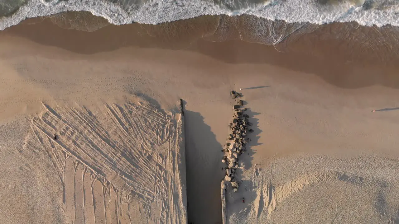 People causing long shadows passing waves coming in on a pristine Leblon beach with the mouth of a drainage canal contrived of partly constructed and partly placed rocks leading to the city lake